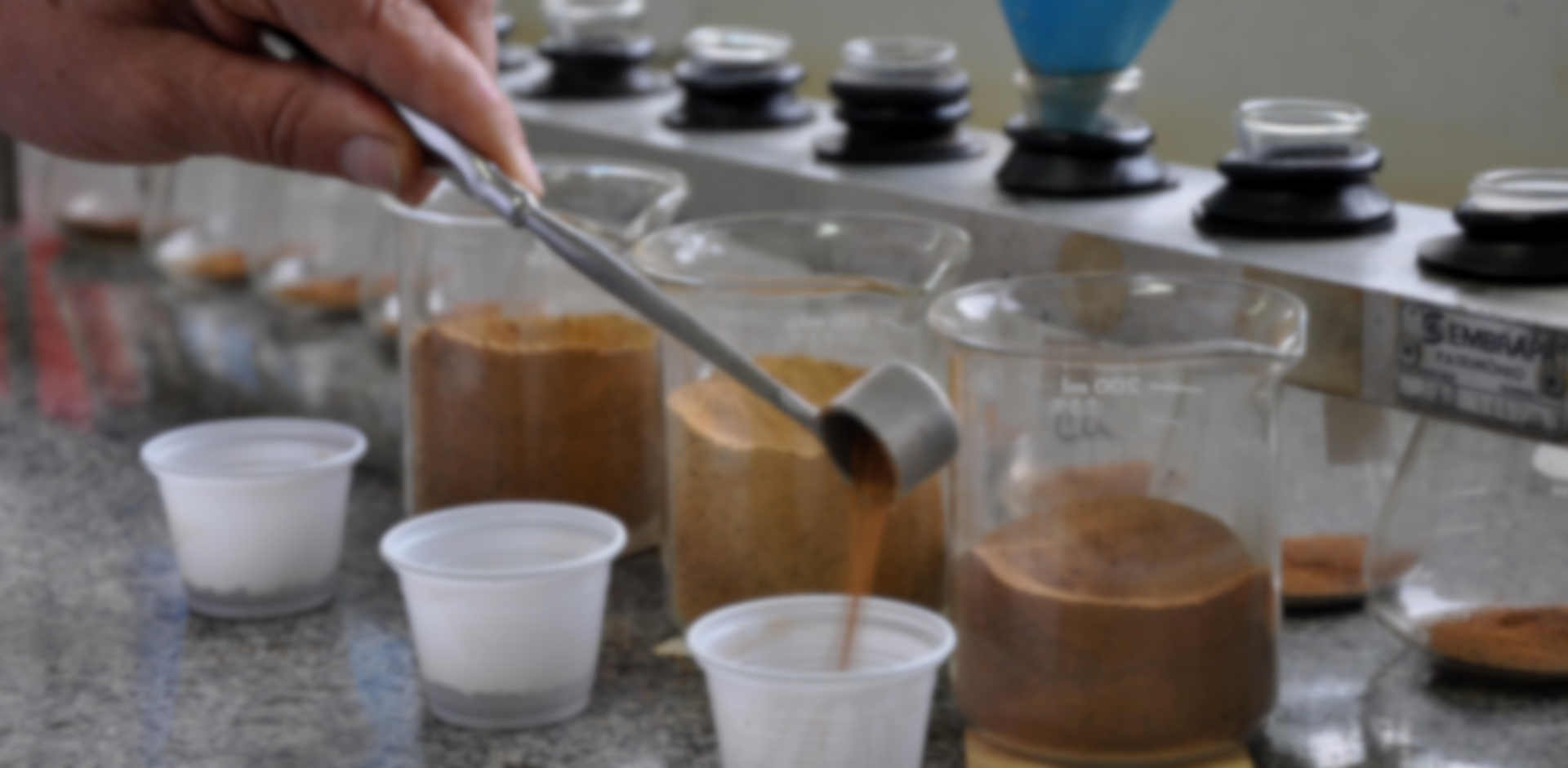 Imagem de uma bancada de laboratório com vários béqueres contendo amostras de solo, e uma mão transferindo solo de um bequer para outro recipiente usando um cachimbo dosador